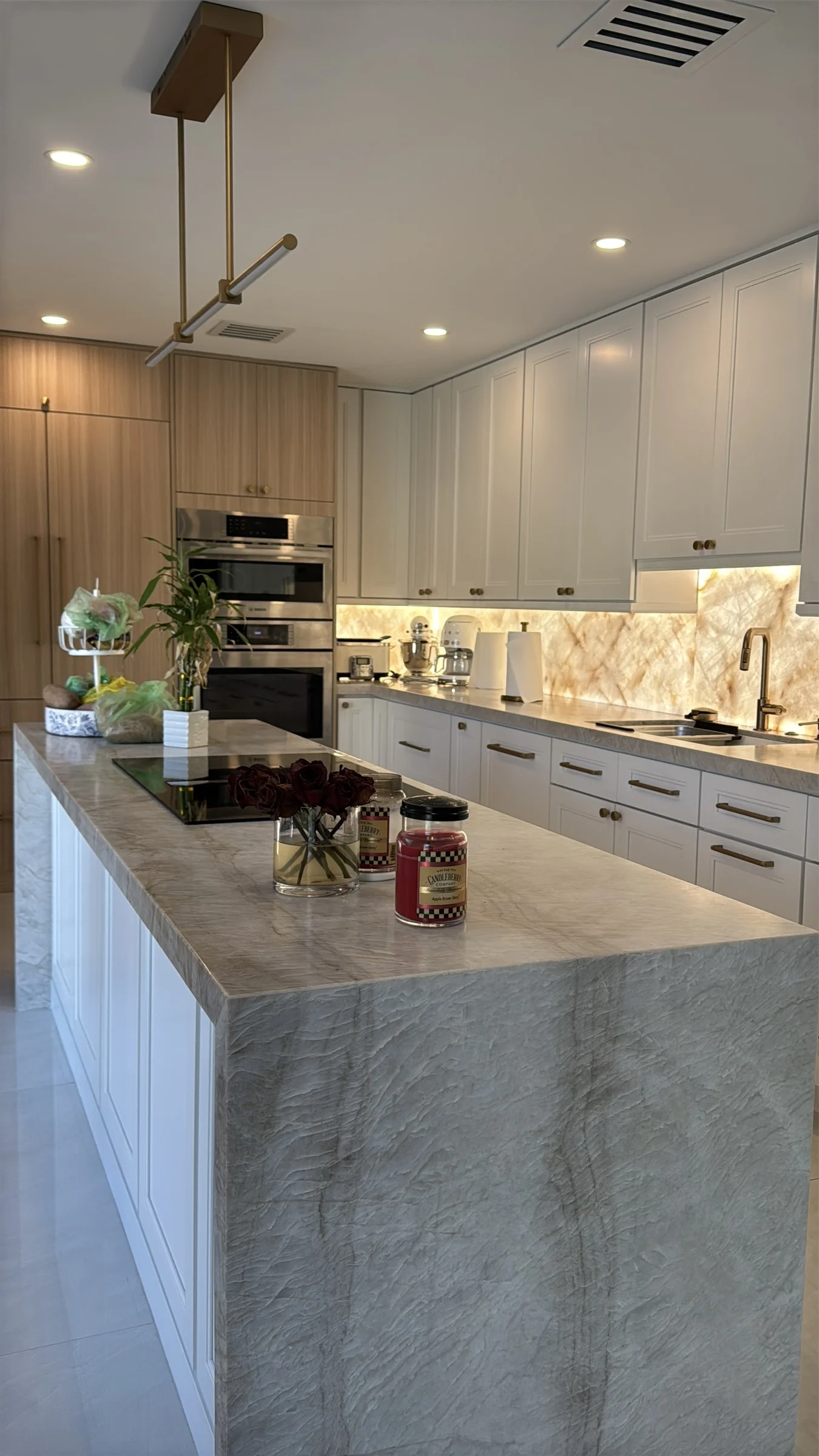 Finished kitchen with white cabinetry, a large stone waterfall island, brass hardware, and an illuminated backsplash